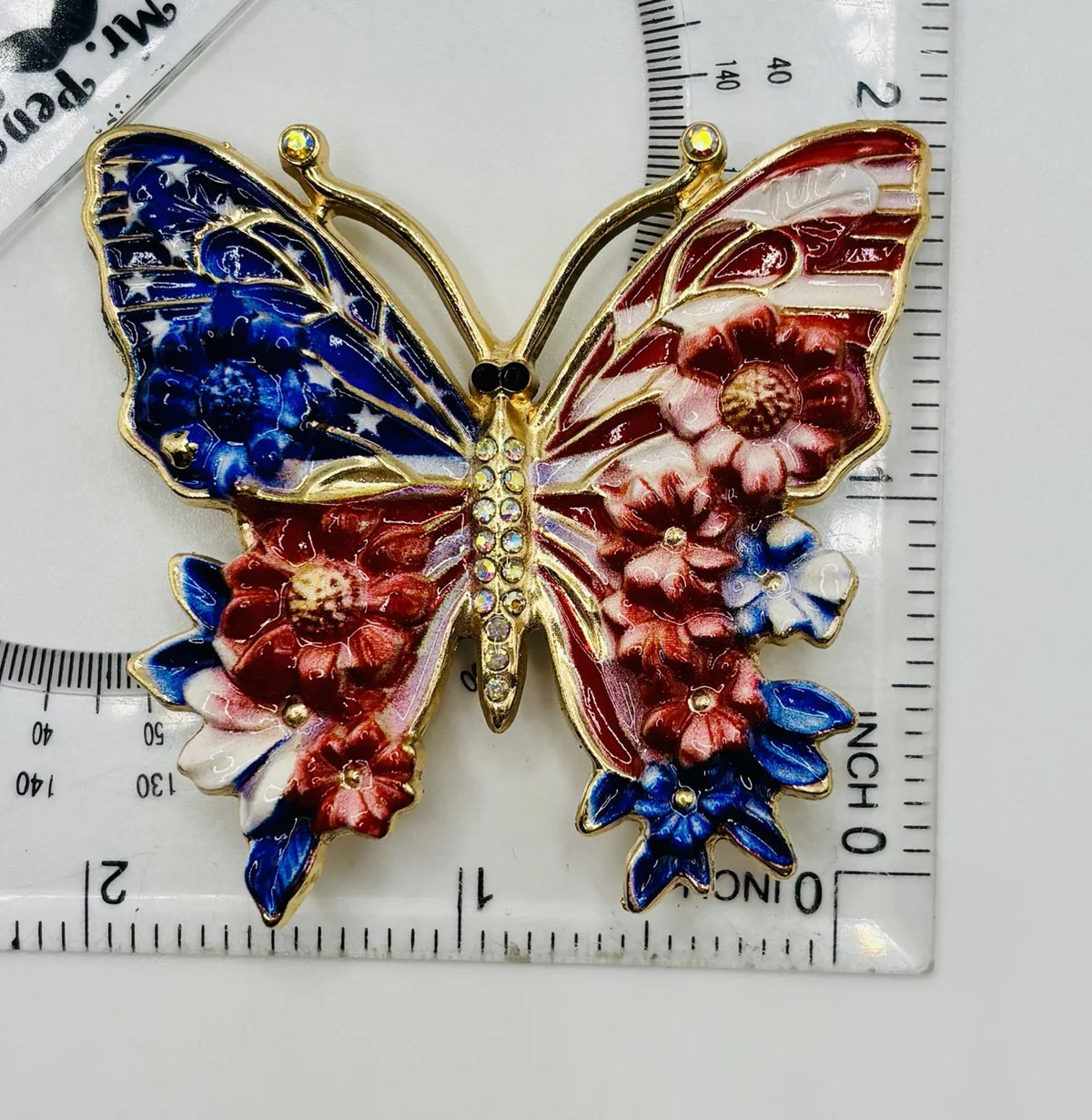 Red white and red white and blue enamel butterfly needle minder beside a ruler for scale