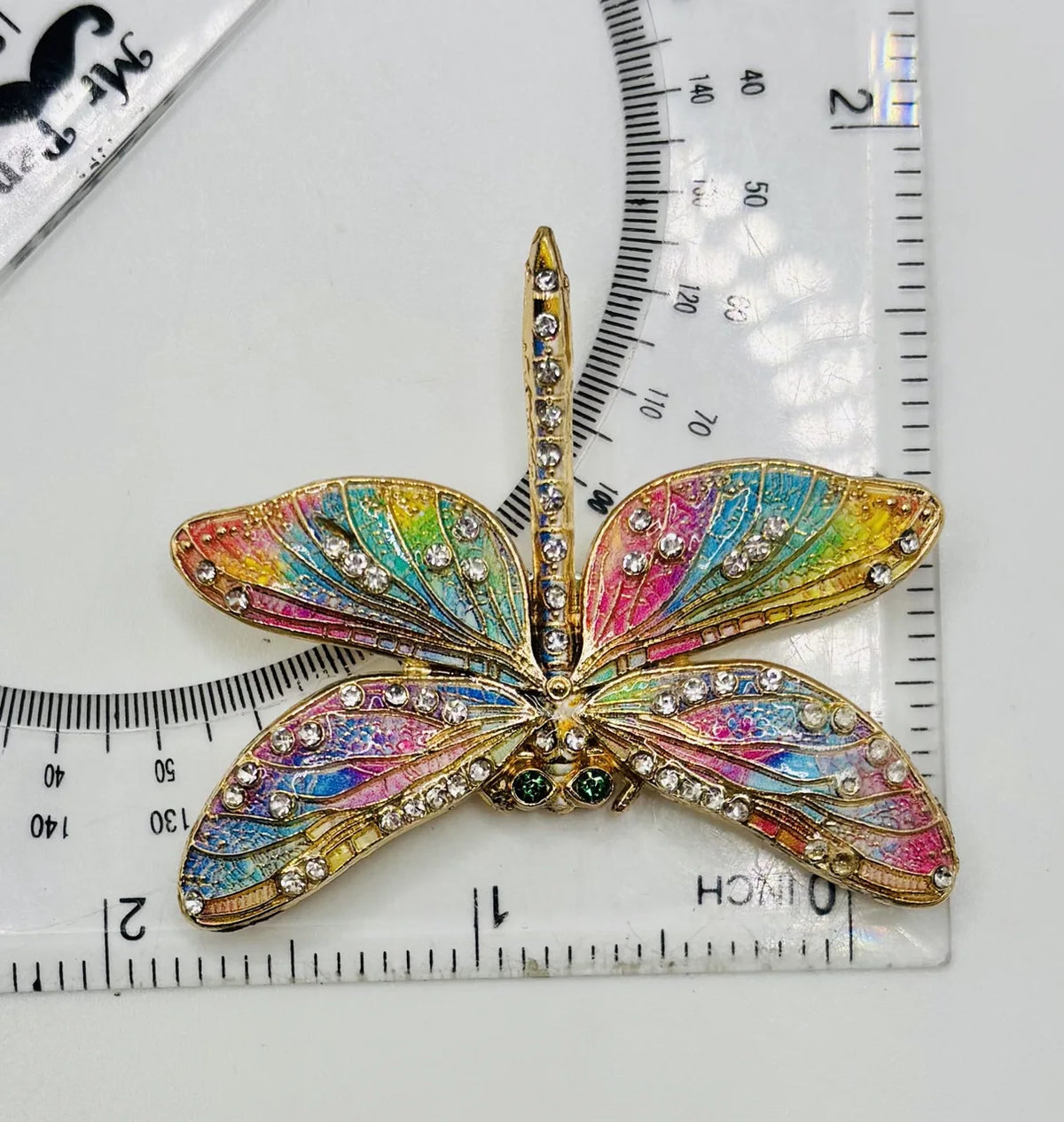 Red white and pastel rainbow enamel crystal dragonfly needle minder beside a ruler for scale