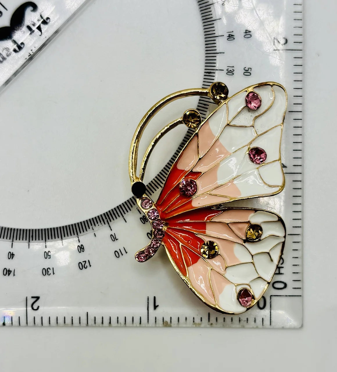 Red white and peach and white enamel and crystal butterfly needle minder beside a ruler for scale